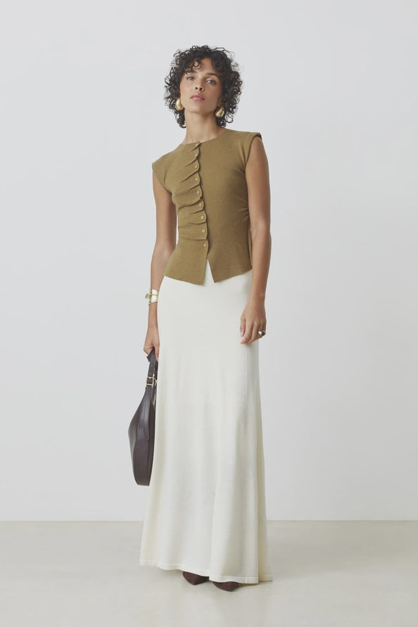 A person with short curly hair and earrings wears the SUNCOO PAGNI S/L button front brown sleeveless jumper and a long white skirt, holding a dark handbag and wearing bracelets, standing against a plain background.
