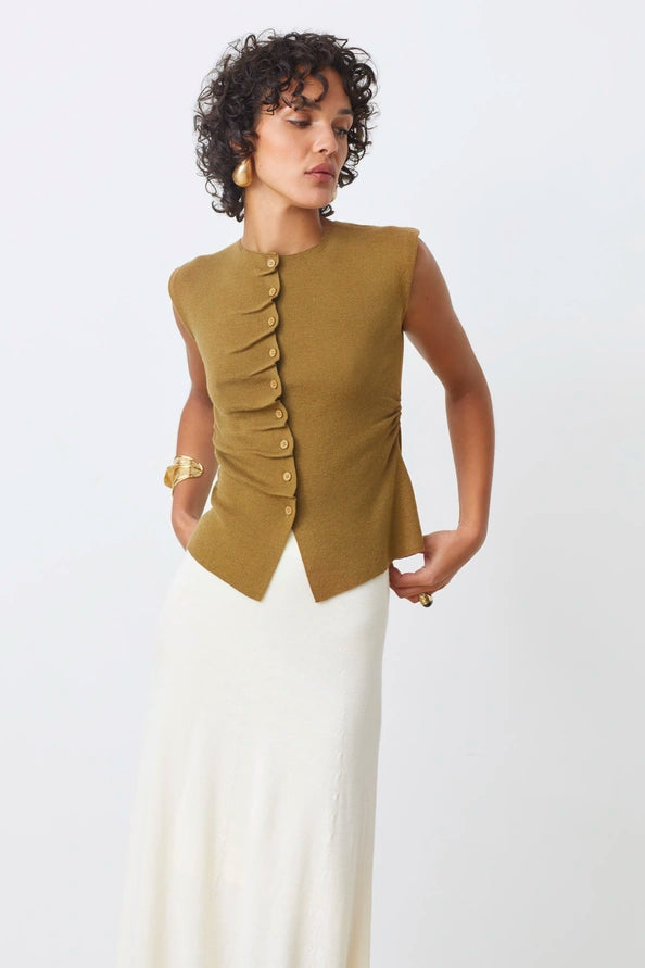 A woman models the SUNCOO PAGNI S/L button front jumper in olive green with an asymmetrical cut, paired with a long white skirt, against a plain white background, gazing slightly to the side.