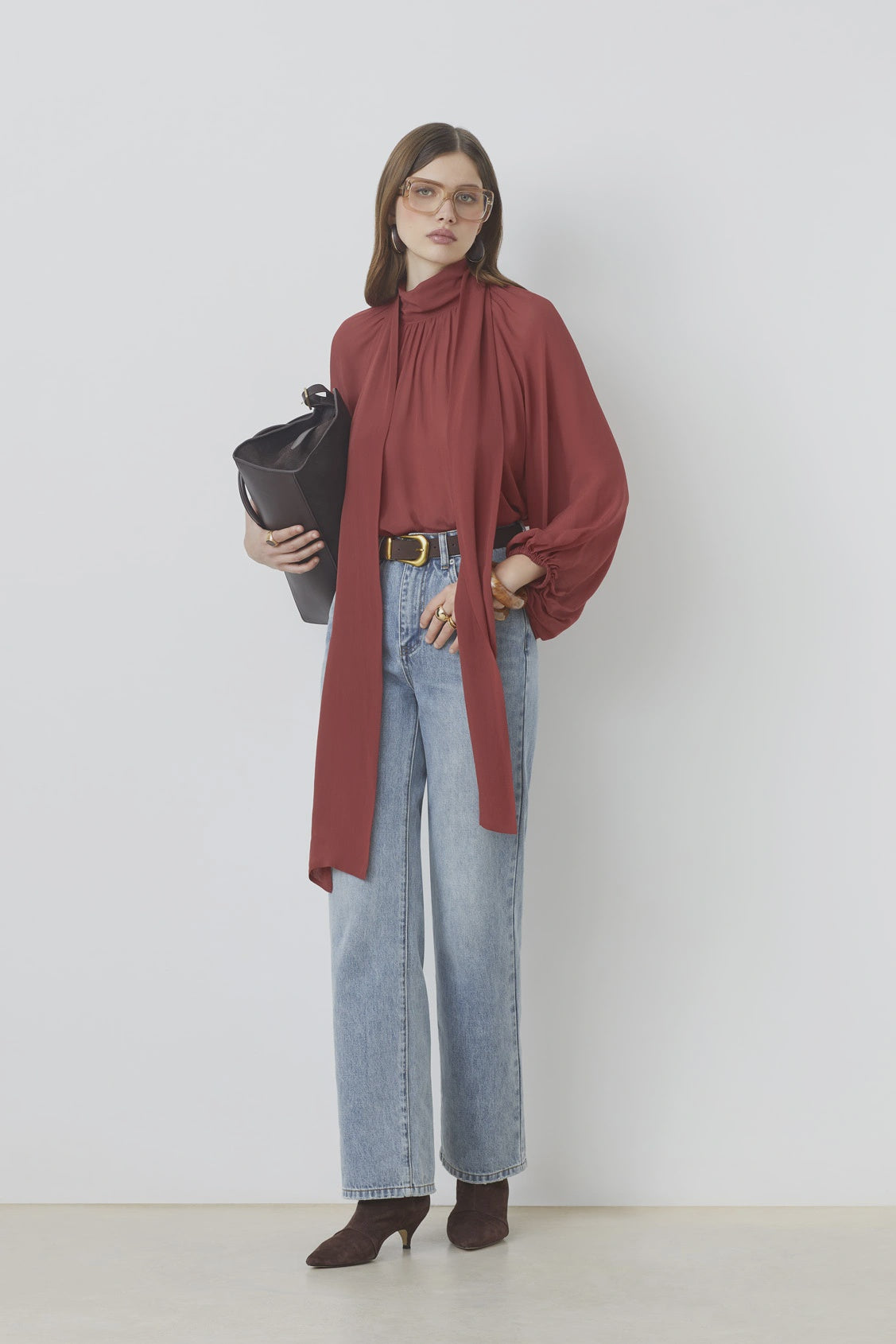 A woman stands against a white wall wearing the SUNCOO LINCOLN blouse with balloon sleeves and a bow collar, light blue high-waisted jeans, brown heeled boots, glasses, and holding a large black bag.