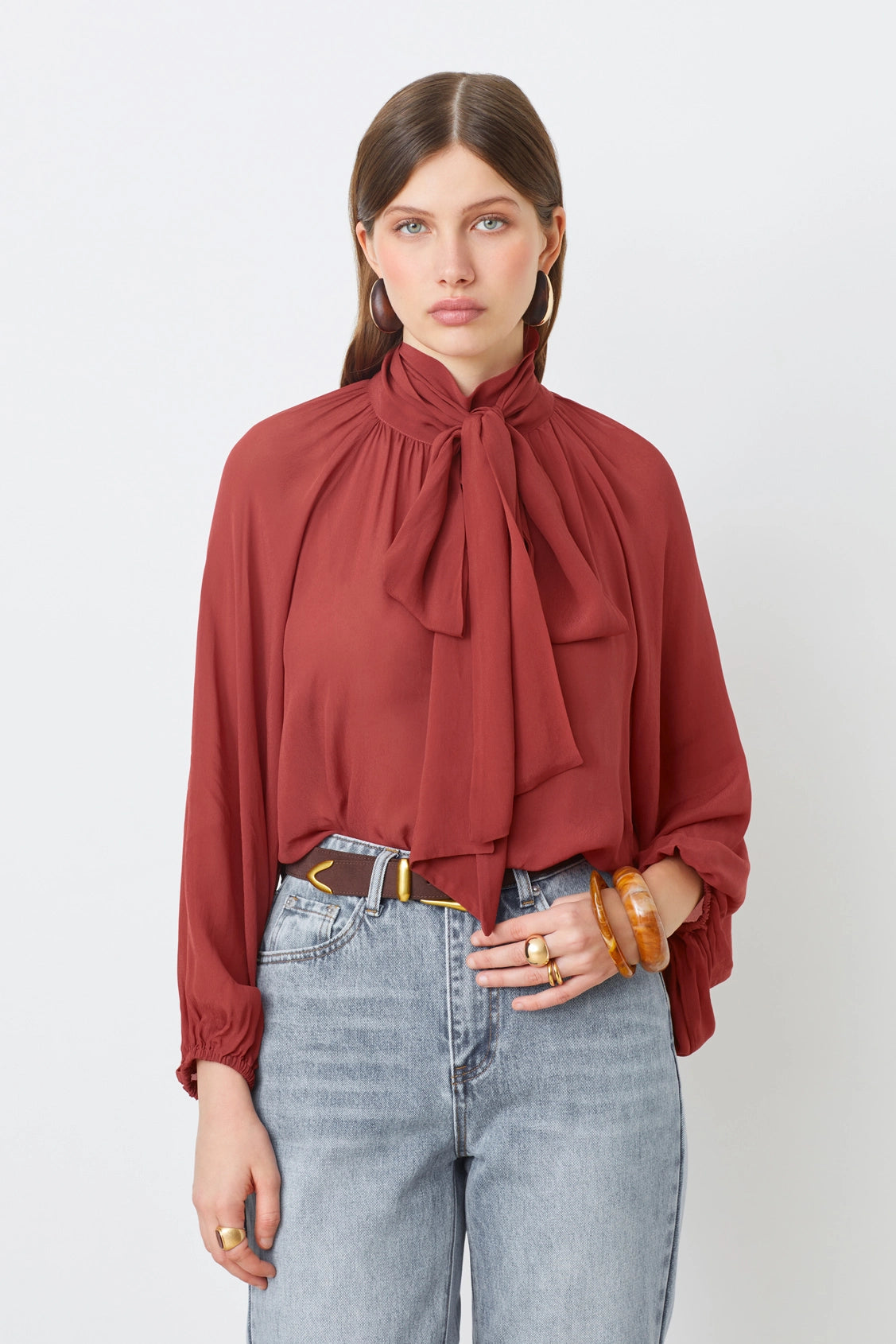 A woman poses against a white background wearing the SUNCOO LINCOLN blouse in rust muslin with a bow collar, paired with high-waisted light blue jeans and accessorized with hoop earrings, rings, and a chunky bracelet.