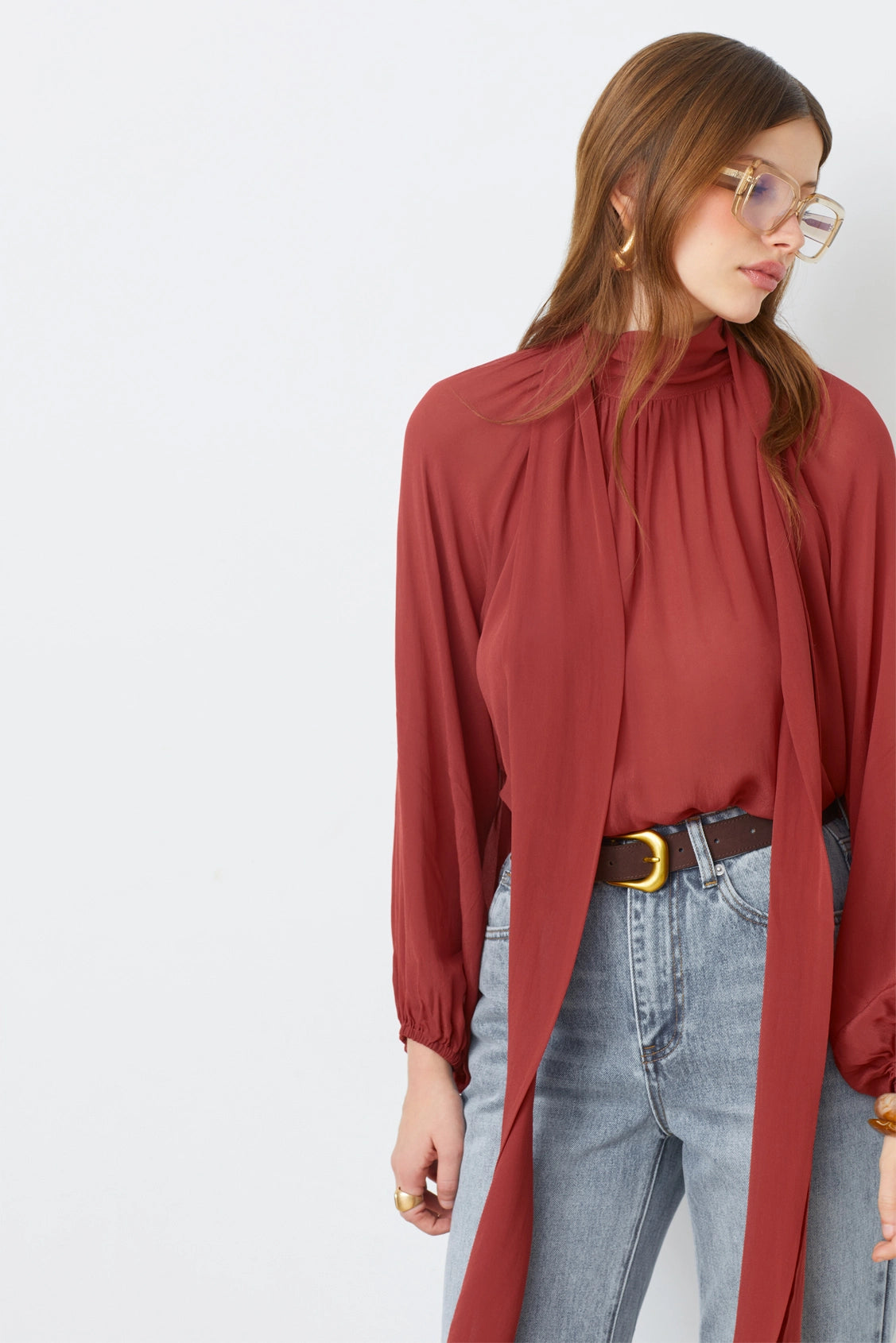 A woman wearing large glasses and the SUNCOO LINCOLN blouse—rust-red muslin with a scarf detail and balloon sleeves—paired with light blue high-waisted jeans and a brown belt, stands against a white background, looking down and to the side.