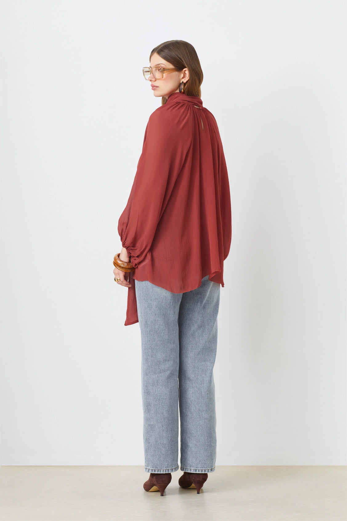 A woman stands facing away against a white background, wearing the SUNCOO LINCOLN blouse with balloon sleeves, light blue jeans, brown heels, and large glasses.