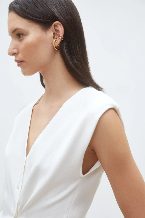 A woman with straight dark hair, wearing the SUNCOO Gilem Vest—a sleeveless white V-neck top with subtle shoulder pads—is shown in profile against a plain light background, accessorized with multiple gold hoop earrings.