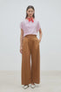 A woman stands against a plain background wearing SUNCOO’s Woven Pantalon Jayden, a pink striped top, white studded shoes, and a red scarf tied at her neck. Her hands are in the pockets of the wide-leg pants with a drawstring waistband.