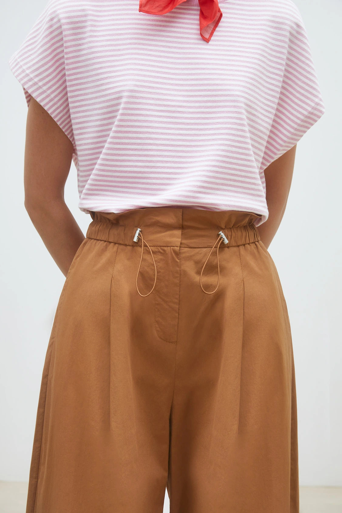 A person stands with their hands behind their back wearing brown cotton poplin Woven Pantalon Jayden by SUNCOO, paired with a pink and white striped short-sleeve top.