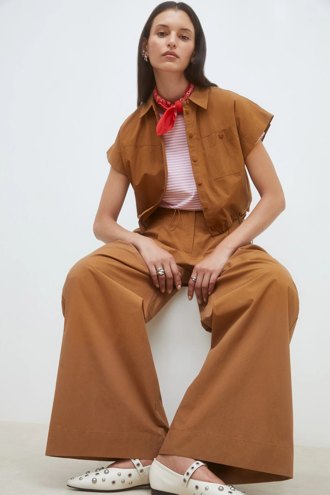 A woman sits on a stool wearing SUNCOO’s Woven Pantalon Jayden wide-leg pants and a brown short-sleeve button-up shirt, accessorized with a red scarf, pink-striped top, white studded shoes, and silver rings as she looks confidently at the camera.
