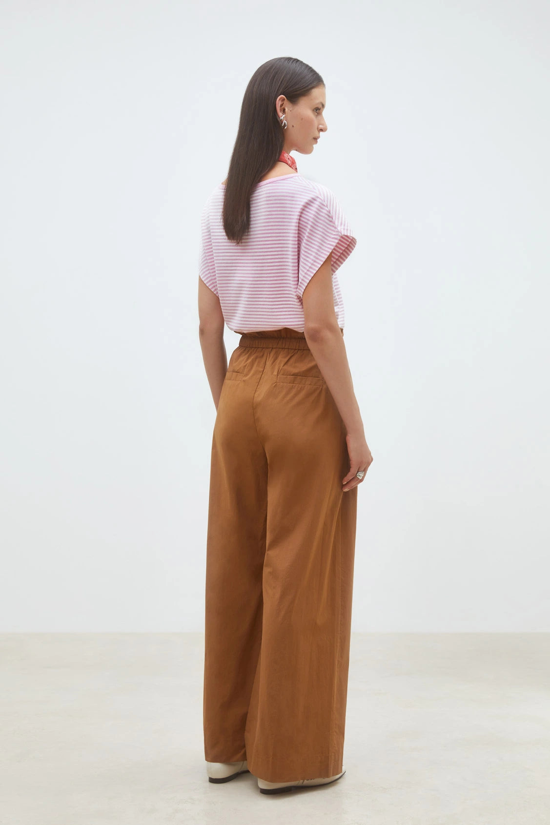 A woman stands indoors with her back to the camera, wearing SUNCOO's Woven Pantalon Jayden in brown organic cotton, paired with a pink and white striped short-sleeve top and cream shoes against a plain white wall.