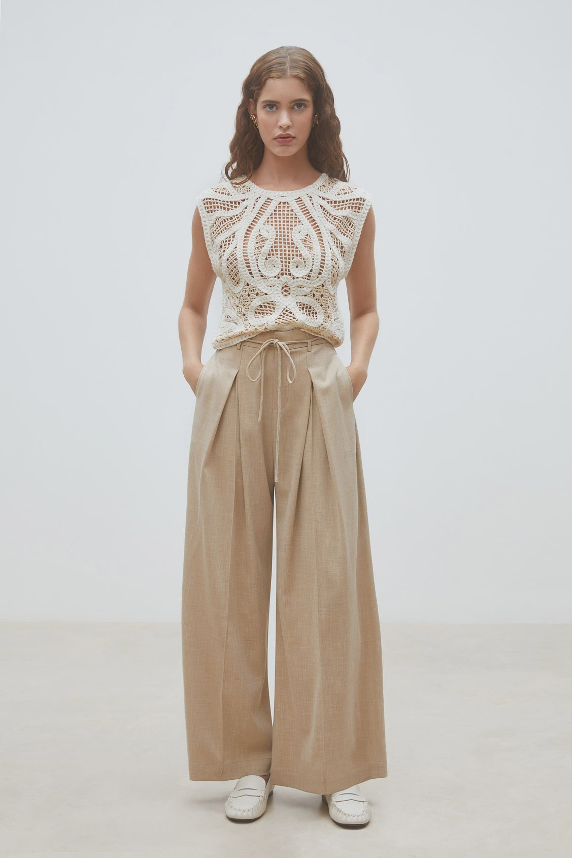 A woman stands against a plain background wearing SUNCOO's Woven Pantalon Jeff—high-waisted darted wide-leg pants with pleats—paired with a sleeveless white crochet top and crisp white shoes, her hands in her pockets.