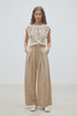 A woman stands against a plain background wearing SUNCOO's Woven Pantalon Jeff—high-waisted darted wide-leg pants with pleats—paired with a sleeveless white crochet top and crisp white shoes, her hands in her pockets.