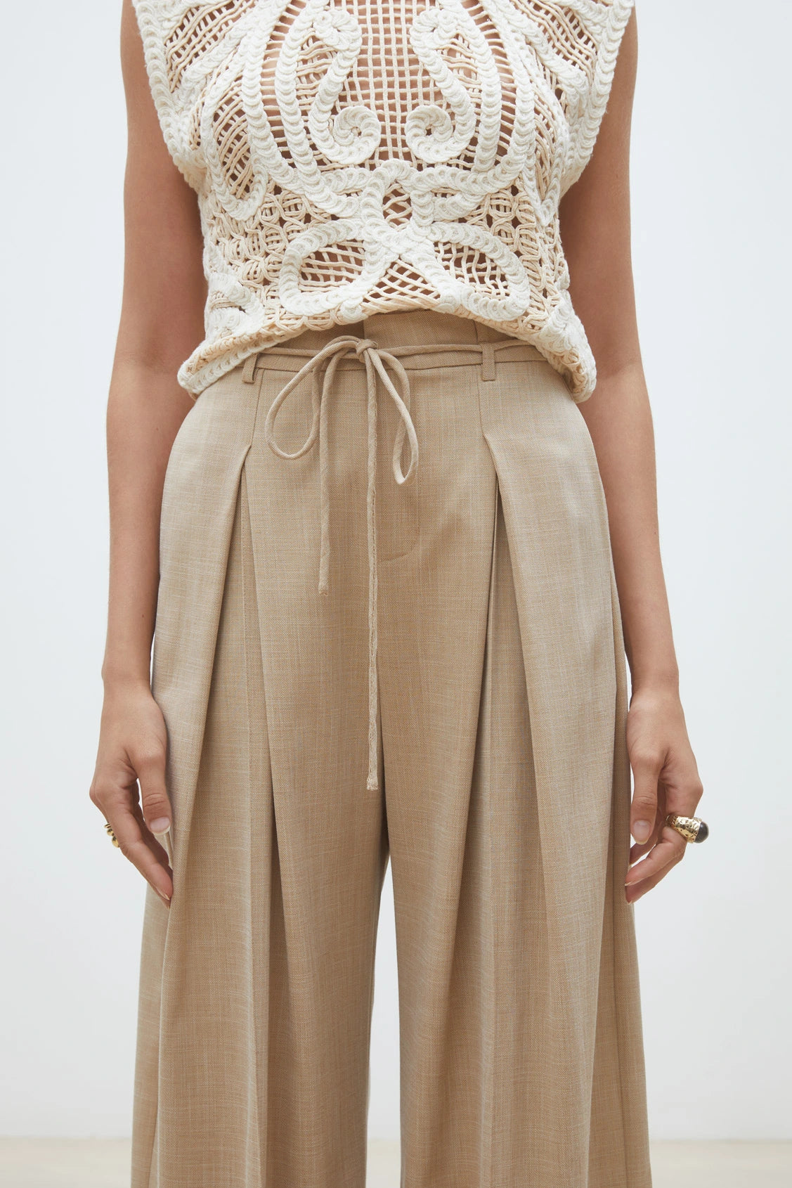 A person models SUNCOO's Woven Pantalon Jeff—high-waisted, wide-leg pants with pleats and a drawstring—paired with a sleeveless, intricately crocheted cream top. Only the torso and upper legs are shown.
