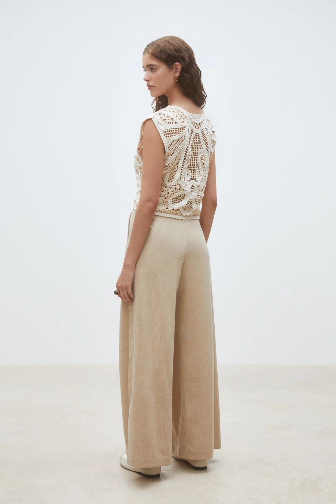 A woman stands in a minimalist, light-toned room, wearing SUNCOO's Woven Pantalon Jeff—high-waisted, wide-leg pants crafted from FSC viscose—paired with a sleeveless cream crochet top.