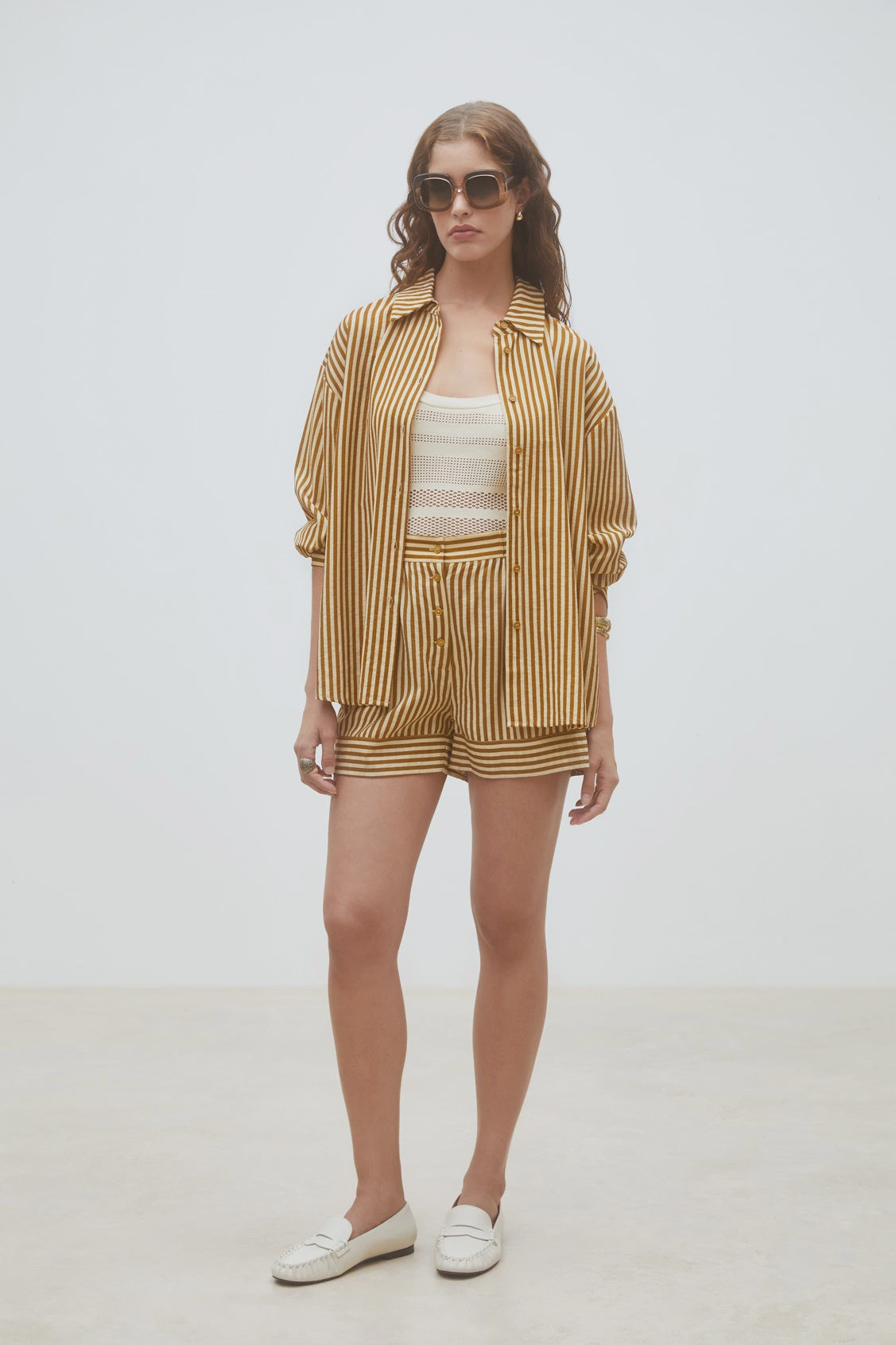 A woman stands against a plain light background wearing SUNCOO's Woven Short Branden, styled with a yellow and white vertically striped shirt, white cropped top, sunglasses, and white loafers.