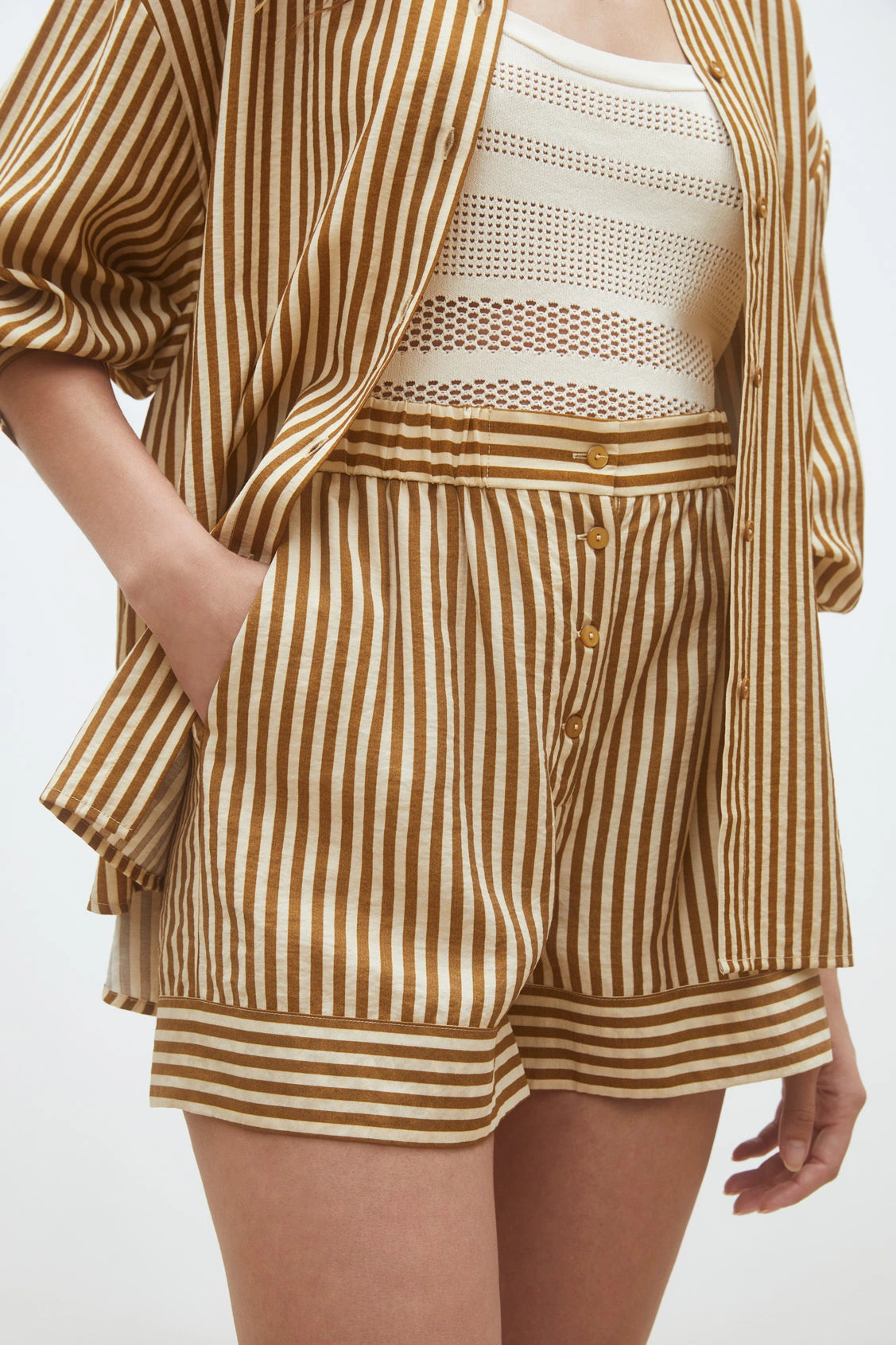 Wearing SUNCOO's Woven Short Branden, a loose brown and white striped button-up, and a white crocheted crop top, the person poses with one hand in their pocket.