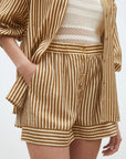 Wearing SUNCOO's Woven Short Branden, a loose brown and white striped button-up, and a white crocheted crop top, the person poses with one hand in their pocket.