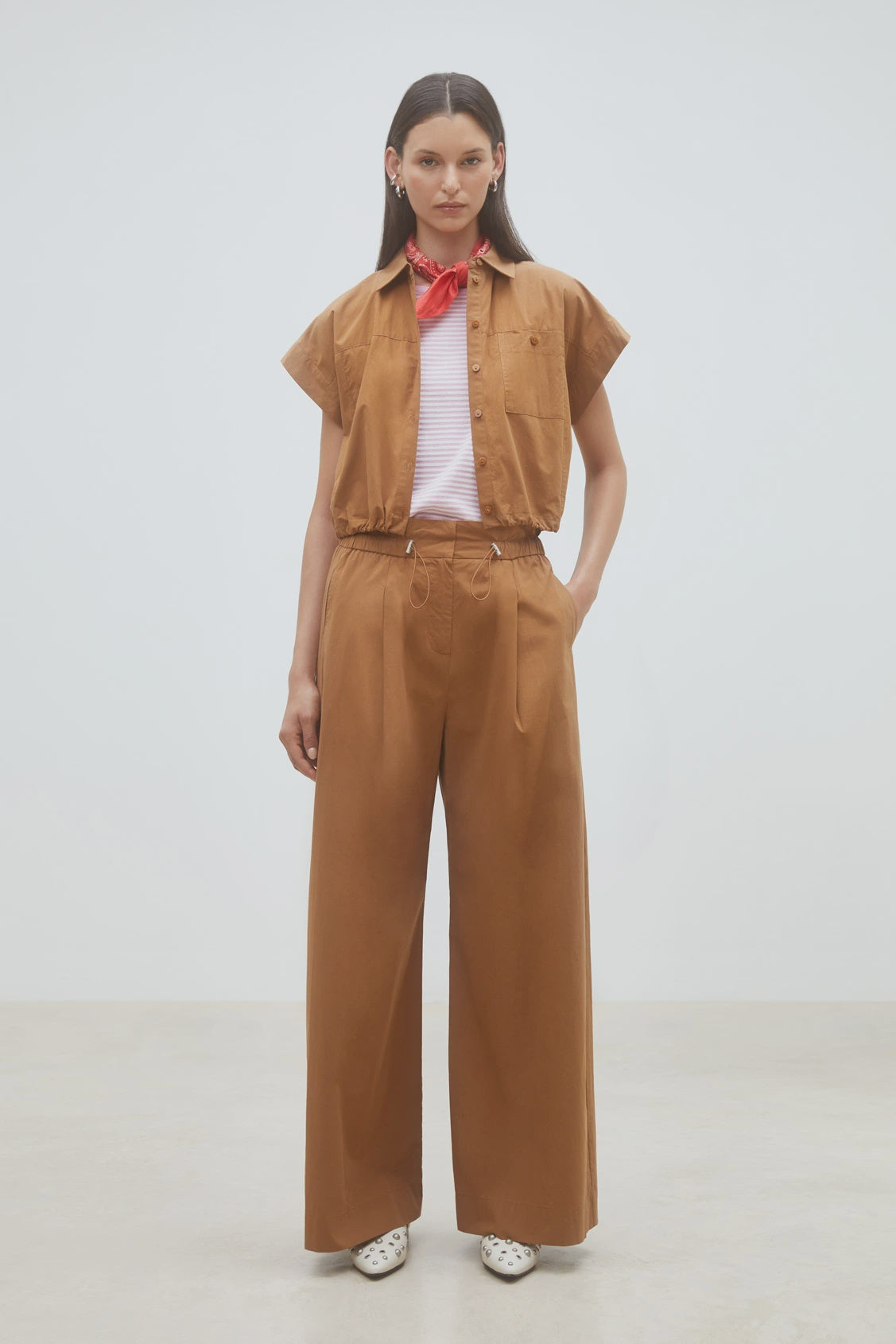 A woman stands against a plain background wearing the SUNCOO Woven Blouse Lorena in brown, matched with wide-leg trousers, a red neck scarf, a pale pink top underneath, and white shoes with black spots. Her hands rest in her pockets.