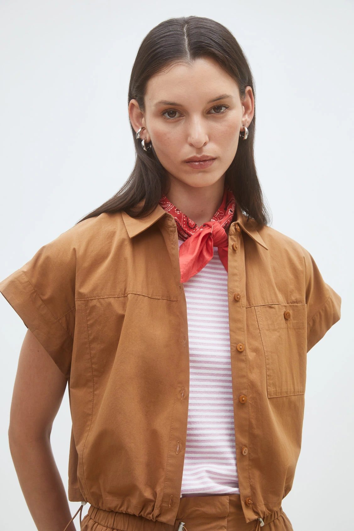 A woman with straight dark hair models the SUNCOO Woven Blouse Lorena, a brown organic cotton short button-down, layered over a light striped top with a red bandana and hoop earrings, standing against a plain light background.