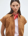 A woman with straight dark hair models the SUNCOO Woven Blouse Lorena, a brown organic cotton short button-down, layered over a light striped top with a red bandana and hoop earrings, standing against a plain light background.