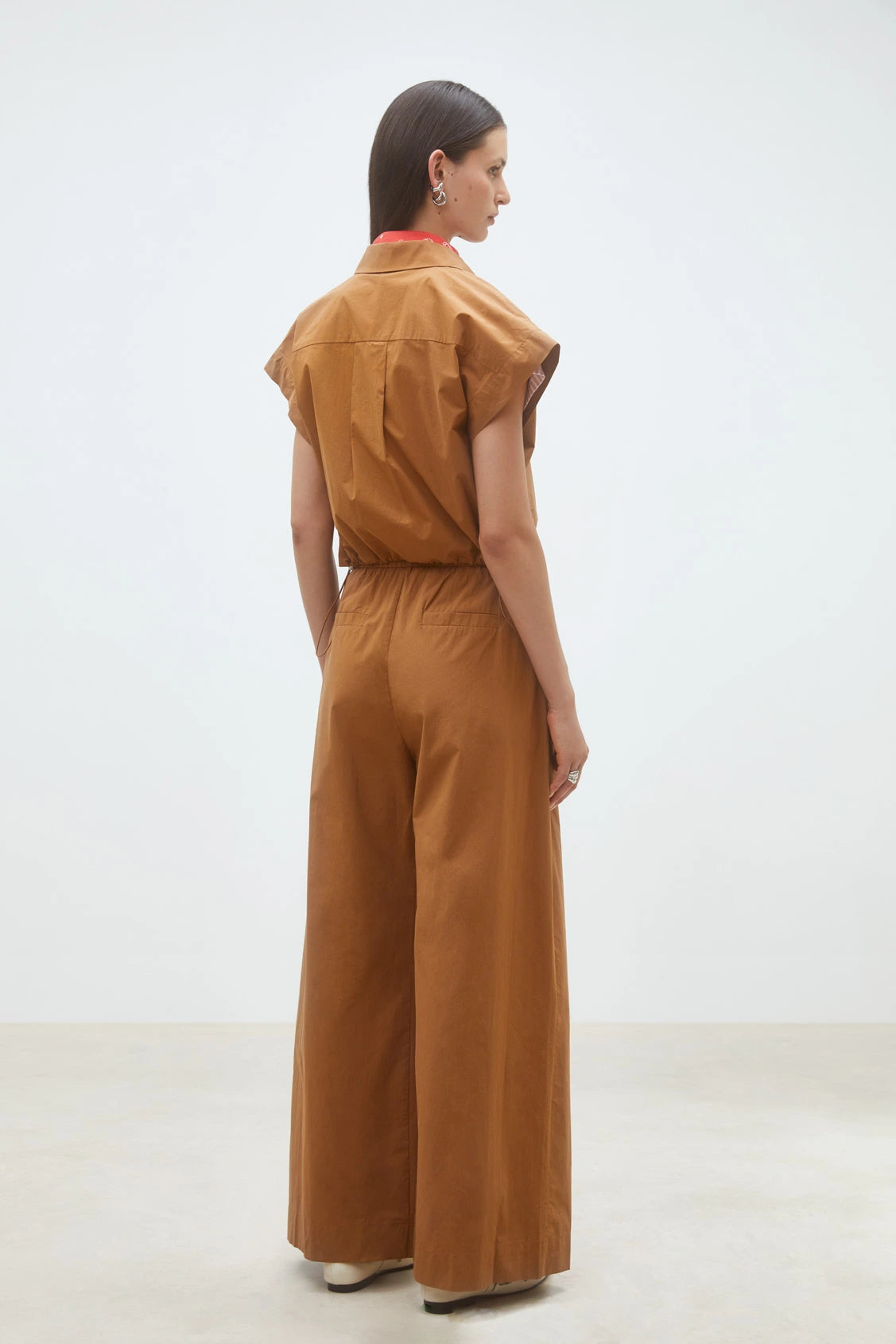 A woman with straight, dark hair stands indoors on a light floor against a white wall, facing away. She wears the SUNCOO Woven Blouse Lorena, a loose-fitting, brown organic cotton top styled as a wide-leg jumpsuit.