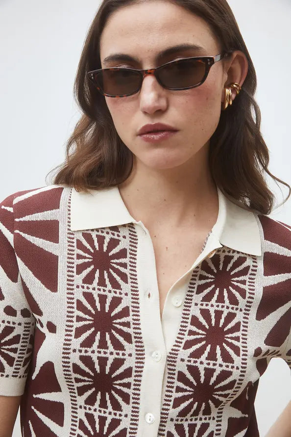 A woman wears sunglasses, gold hoop earrings, and the SUNCOO Gabera Shirt—a jacquard knit cardigan with a white collar and maroon geometric designs—standing against a plain light background.