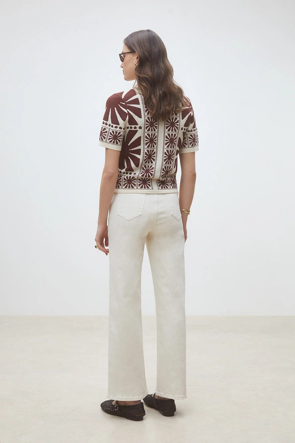 A person stands facing away, wearing the SUNCOO Gabera Shirt, cream wide-leg pants, dark shoes, and sunglasses. The background is plain and light-colored.