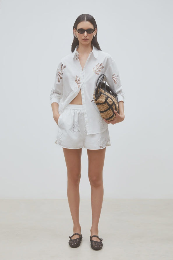 A woman stands against a plain background wearing SUNCOO's Woven Short Bosco with an elastic waistband and a white button-up shirt. She accessorizes with dark sunglasses, black shoes, and a woven straw handbag.