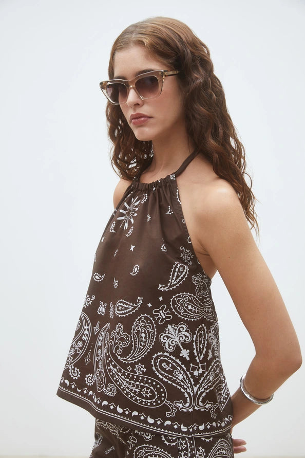 A woman with wavy brown hair wears sunglasses and the SUNCOO Woven Blouse Looka, featuring a white paisley print and halter neck, plus a silver cuff bracelet, standing against a plain white background.