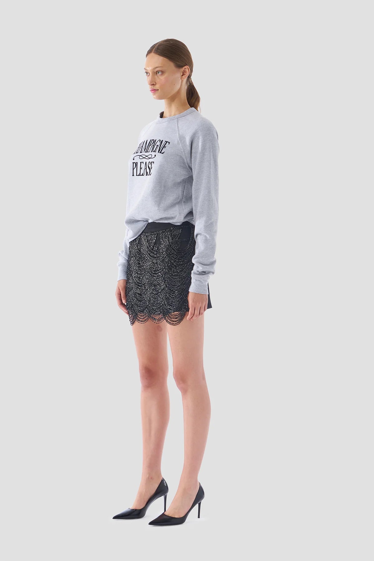 A woman stands against a gray background wearing the "Champagne Please Sweater" by Le Superbe, featuring a handcrafted font. She pairs it with a black lace skirt and elegant black high heels, exuding confidence and style.