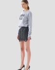 A woman stands against a gray background wearing the "Champagne Please Sweater" by Le Superbe, featuring a handcrafted font. She pairs it with a black lace skirt and elegant black high heels, exuding confidence and style.