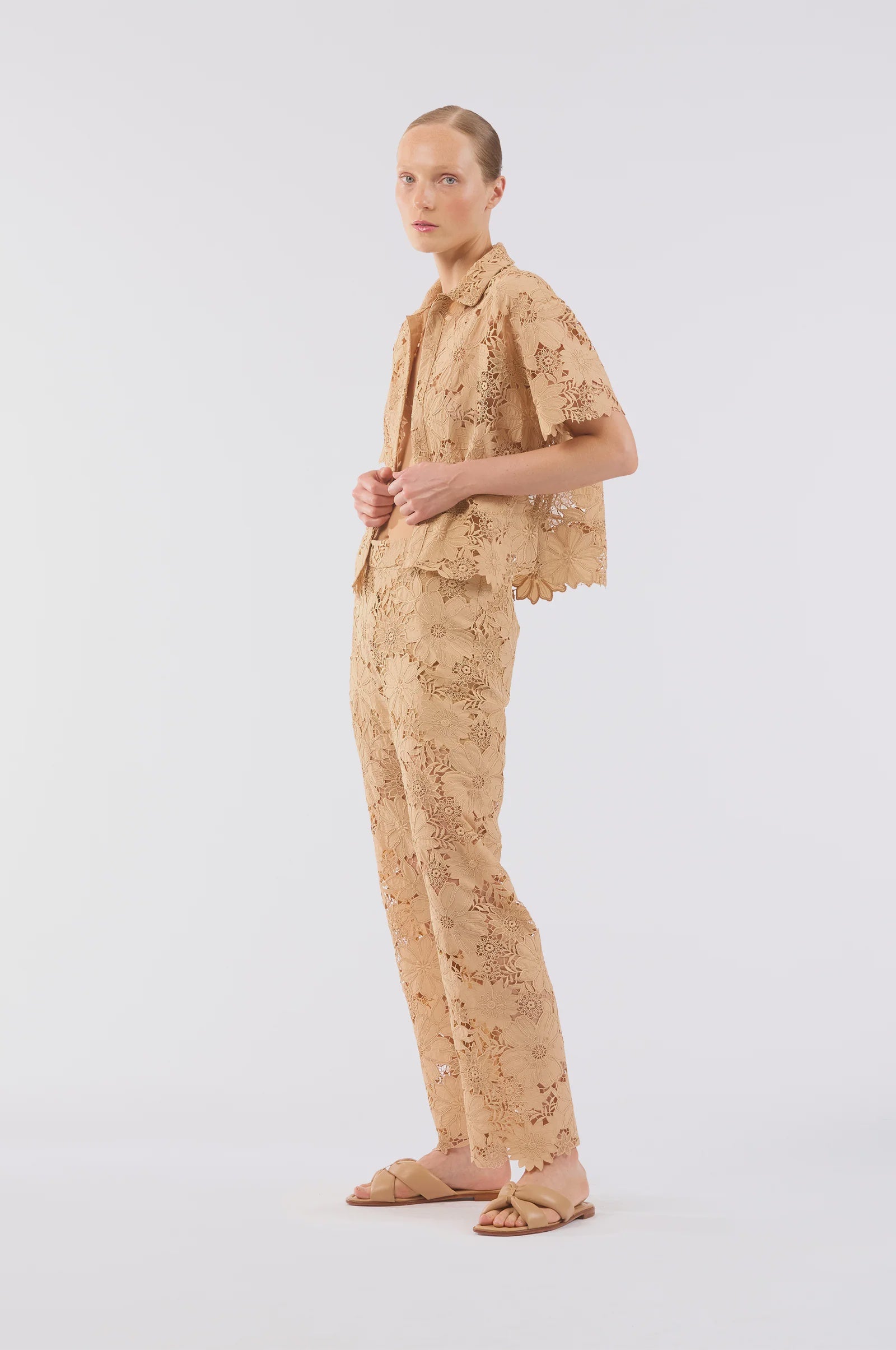 A person wears the Le Superbe Suntan Camp Shirt—light brown, floral lace with scalloped edges—paired with matching pants and tan slide sandals. Their light hair is pulled back and their expression is neutral against a white background.