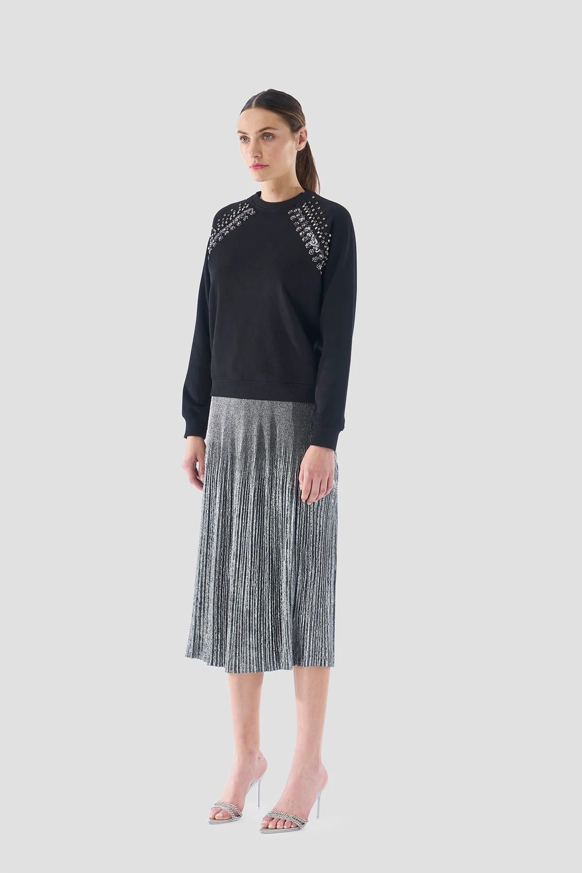 A woman exudes elegance and glamour against a plain backdrop, dressed in a black sweater with embellished shoulders and Le Superbe's Foil Pleated Skirt. She complements the ensemble with silver high heels, while her long hair is gracefully tied back.