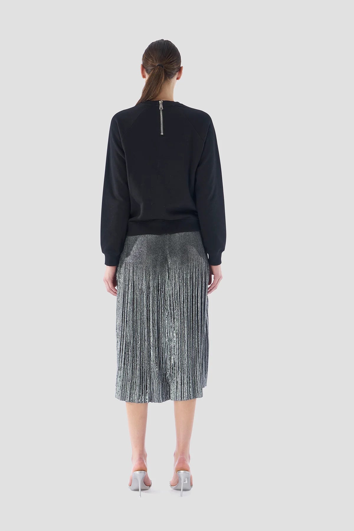 With long hair cascading from the back, they exude elegance in a black sweater featuring a zipper. Paired with Le Superbe's Foil Pleated Skirt and set against a plain gray background, their look is completed with glamorous transparent high heels.