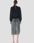 With long hair cascading from the back, they exude elegance in a black sweater featuring a zipper. Paired with Le Superbe's Foil Pleated Skirt and set against a plain gray background, their look is completed with glamorous transparent high heels.