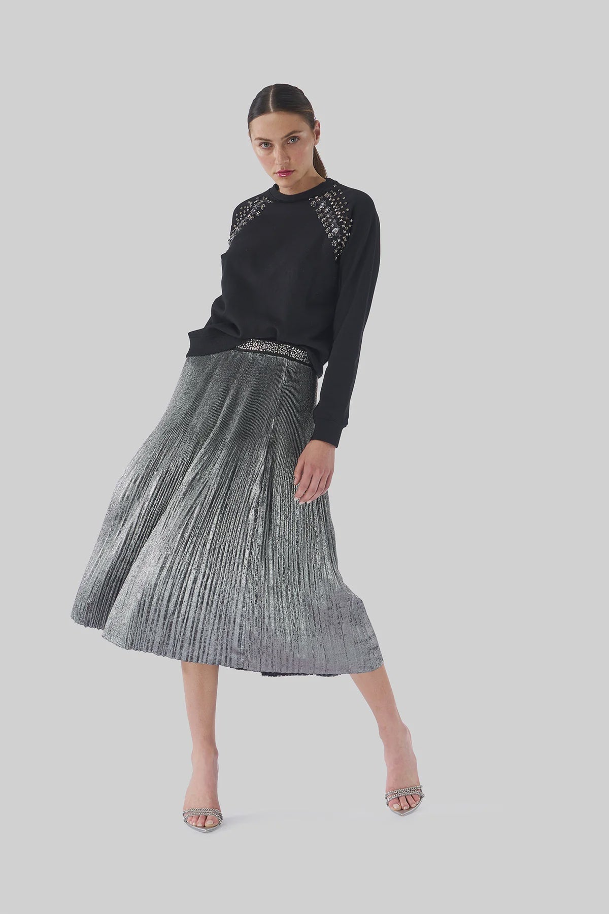 A woman exudes elegance in a black sweater featuring lace shoulder details and the Le Superbe Foil Pleated Skirt as she poses against a plain background. Her look is completed with high heels and her hair tied back, capturing a timeless sense of glamour.