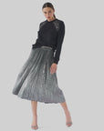 A woman exudes elegance in a black sweater featuring lace shoulder details and the Le Superbe Foil Pleated Skirt as she poses against a plain background. Her look is completed with high heels and her hair tied back, capturing a timeless sense of glamour.