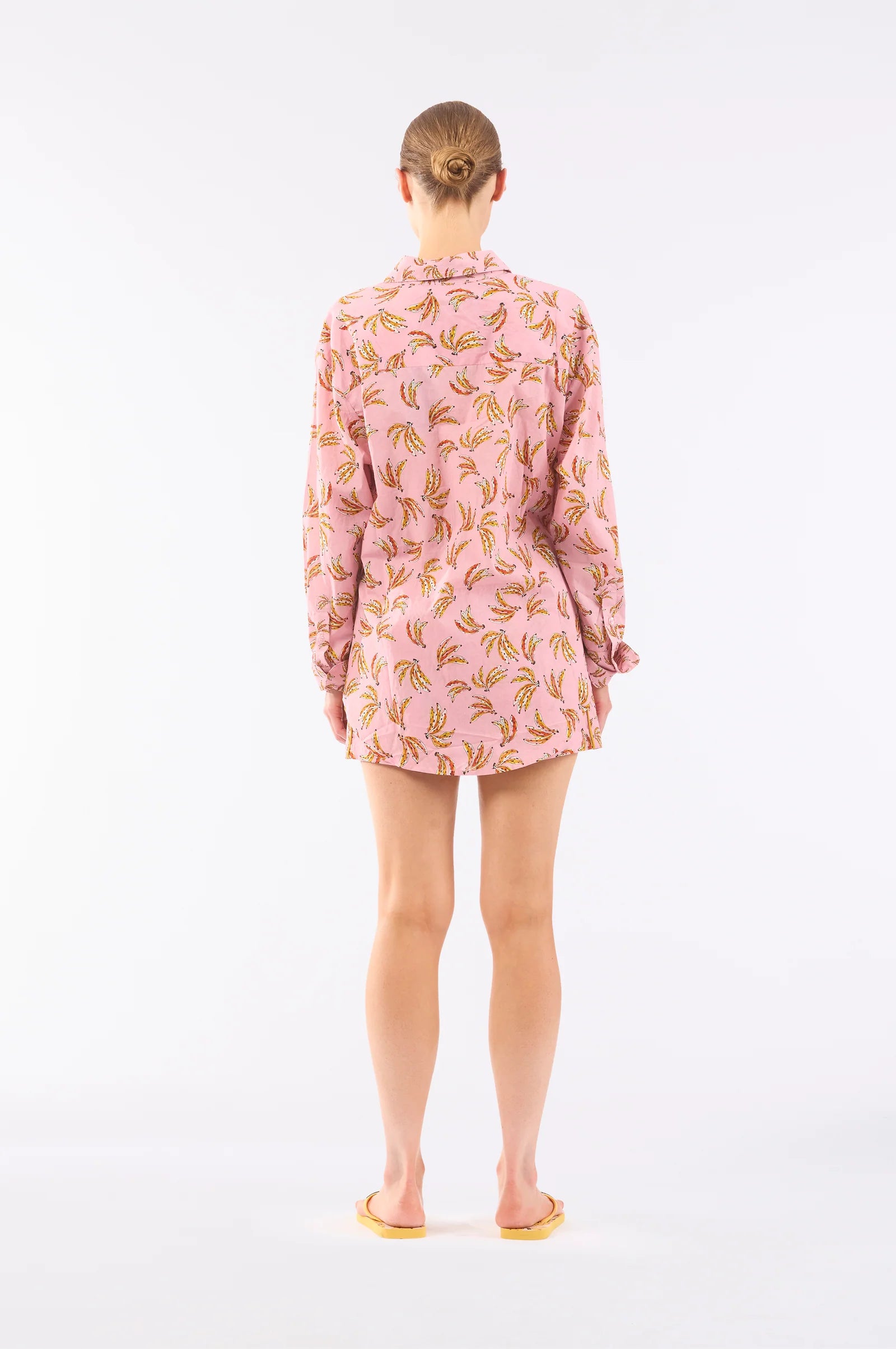 Facing away from the camera, a person wears Le Superbe's Bananas Over You shirt, a pink long-sleeve woven poplin design with a leafy pattern and button closure. Their hair is tied up, and they stand in slippers against a plain white backdrop.