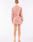 A person with a bun is wearing Le Superbe’s Bananas Surf Short, a long-sleeve pink dress featuring a subtle banana print, standing against a plain white background and paired with yellow flat sandals.