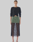 A person is wearing the Twisted Sister Sweater by Le Superbe, which features a soft knit in black, paired with a green mini skirt layered over a long black lace skirt and black heels. They are standing against a plain light gray background.