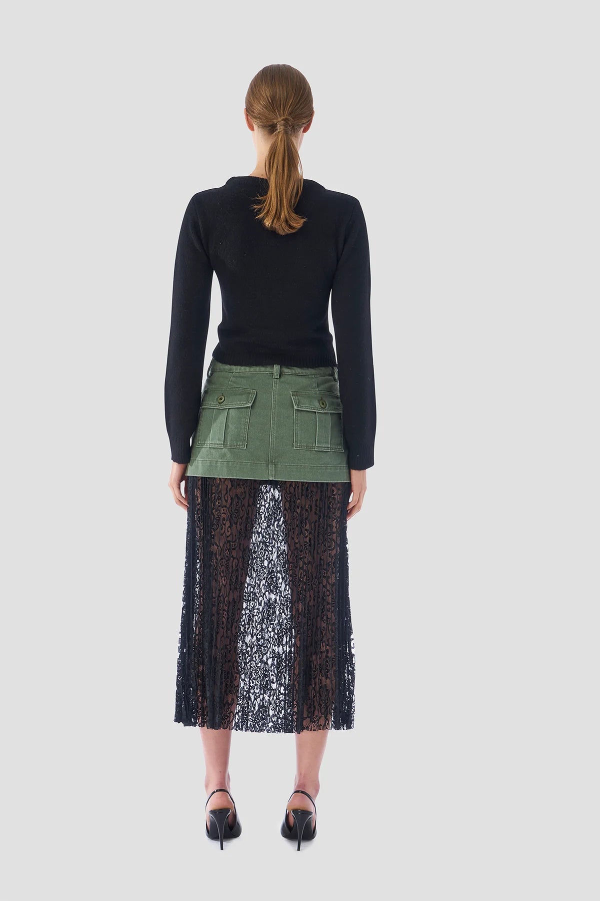 A person with long hair stands facing away, showcasing a sleek black Twisted Sister Sweater by Le Superbe that exudes a modern flair, paired with a green skirt and a flowing black lace overlay. They complete the chic ensemble with black heels against a plain white background.