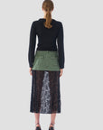 A person with long hair stands facing away, showcasing a sleek black Twisted Sister Sweater by Le Superbe that exudes a modern flair, paired with a green skirt and a flowing black lace overlay. They complete the chic ensemble with black heels against a plain white background.