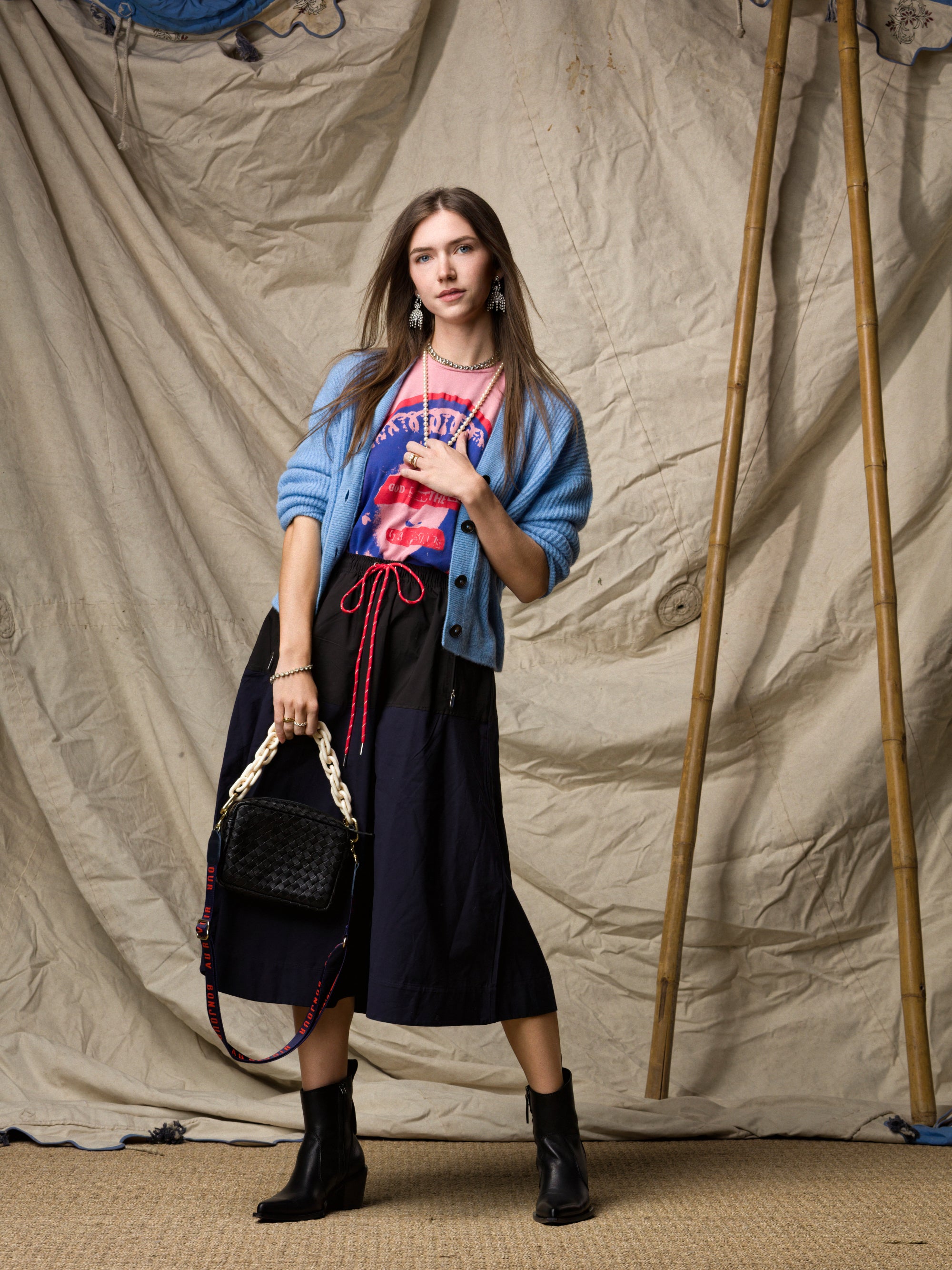 A woman poses before a canvas backdrop wearing a light blue cardigan, pink graphic tee, black ankle boots, a black braided-strap handbag, and the Le Vintage Sport Skirt by Ping Pong Surf Club.