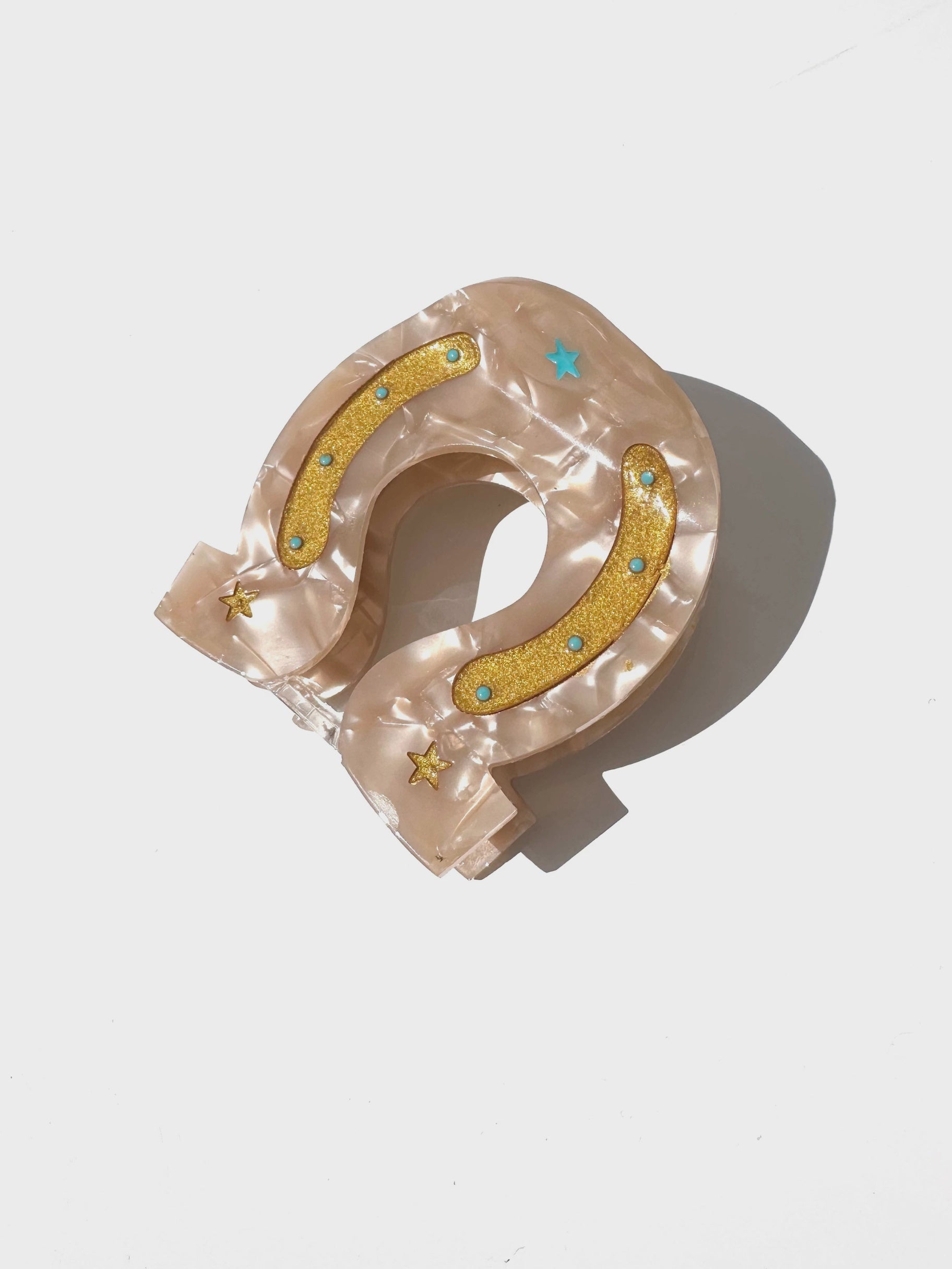 The Fashion Hair Clip by Faire is a beige, pearlescent horseshoe-shaped accessory made from eco-friendly cellulose acetate, featuring yellow glitter accents, blue rhinestones, and star details, displayed on a white surface.