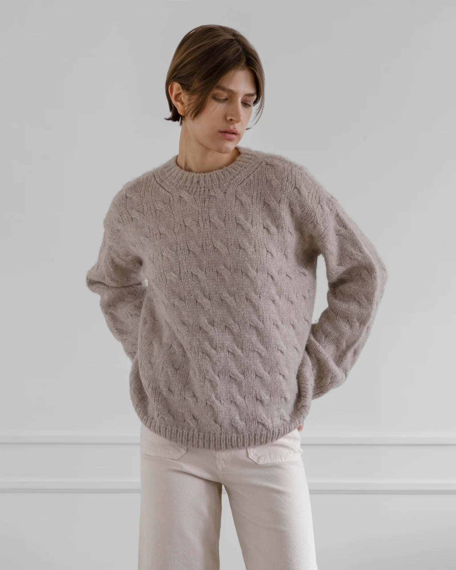 A person with short brown hair wears the LATIERRA Jolie Sweater, a loose, classic fit cable knit in light brown, paired with off-white pants, standing against a plain light gray wall and looking down to the side.