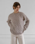 A person with short brown hair wears the LATIERRA Jolie Sweater, a loose, classic fit cable knit in light brown, paired with off-white pants, standing against a plain light gray wall and looking down to the side.