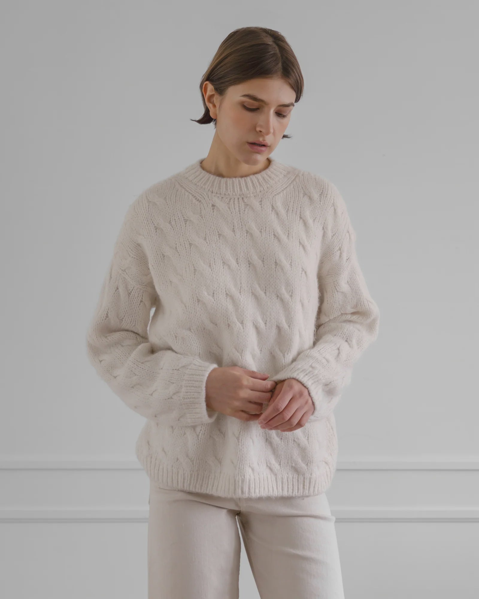 Wearing the LATIERRA Jolie Sweater in cream, a person with short brown hair and light pants stands against a plain light gray background, looking down with hands loosely clasped.