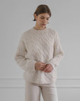 Wearing the LATIERRA Jolie Sweater in cream, a person with short brown hair and light pants stands against a plain light gray background, looking down with hands loosely clasped.