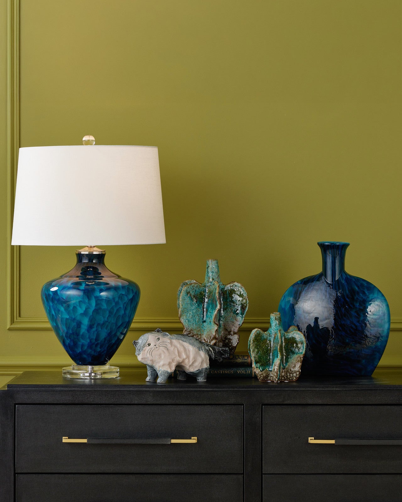 A dark dresser with gold handles displays the Currey & Company, Inc. Siamese Cat sculpture, along with a blue glass lamp, two green vases, and a blue vase, all set against an olive green wall.