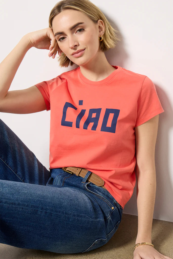 A young woman in the Clare Vivier Classic Tee CIAO—coral, short-sleeved, with bold blue lettering—pairs it with blue jeans and a tan belt. Made of lightweight cotton, she sits on the floor, smiling softly with her head resting on one hand.