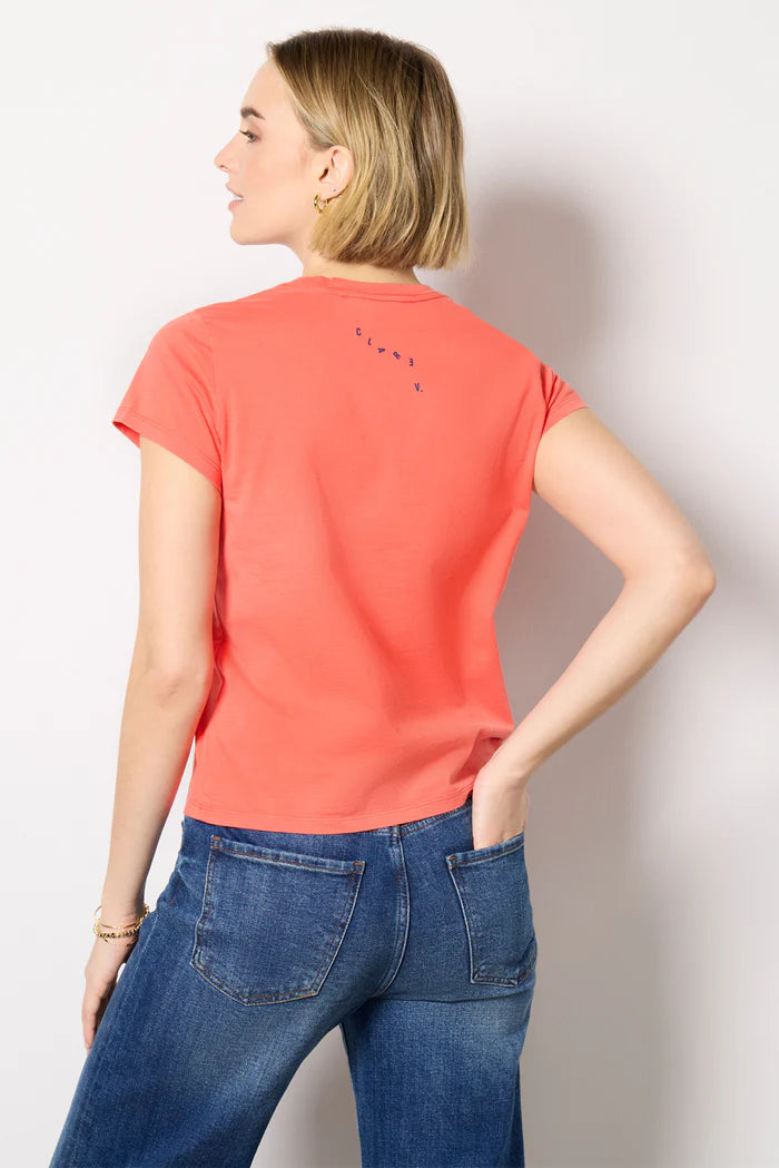 A woman with short blonde hair stands with her back to the camera, one hand in her pocket, wearing Clare Vivier's Classic Tee CIAO in coral and blue jeans against a plain white background.