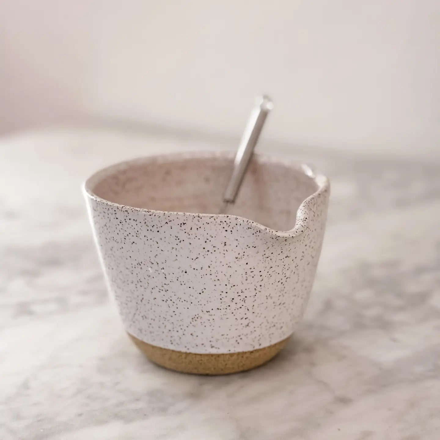 The Faire Ceramic Bowl w/ Whisk, a unique handmade piece with a small spout, sits speckled on marble, accompanied by its metal whisk.