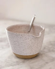 The Faire Ceramic Bowl w/ Whisk, a unique handmade piece with a small spout, sits speckled on marble, accompanied by its metal whisk.